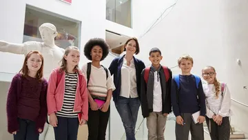 Ein Erwachsener und vier Kinder sitzen an einem Tisch und arbeiten an einem Projekt.  Sie tragen bunte Kleidung. Der Raum ist hell mit Fenstern. Auf dem Tisch liegen Notizbücher.