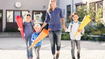 Eine Mutter geht mit drei Kindern vor einer Schule. Die Kinder tragen Rucksäcke und bunte Schultüten (rot, orange, gelb). Alle lächeln und sind lässig gekleidet. Die Schule hat große Fenster und eine Uhr. Die Sonne scheint.