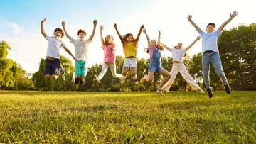 Ein Kind mit langen Haaren holt lächelnd ein Buch aus einem Regal. Es trägt ein beiges Kleid und steht auf einem grünen Teppich. Im Hintergrund gibt es Kissen und Bücher.