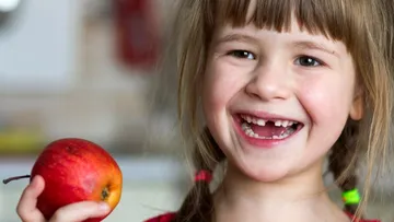 Ein fröhliches Mädchen mit fehlenden Zähnen hält einen Apfel.