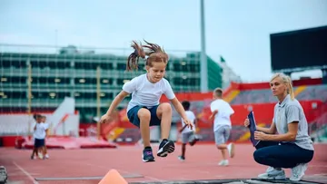 Ein Mädchen macht Weitsprung, während die Trainerin zusieht.
