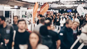 Ein geschäftiges Bild einer Menschenmenge auf einer Messe. Die Leute sind unscharf und tragen dunkle Kleidung. Über ihnen hängen orangefarbene Fahnen mit Logos. Im Hintergrund sieht man Glaswände und Bildschirme.