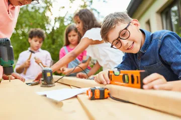 Kinder bauen in einem sonnigen Garten mit Holz- Ein Junge misst mit einer Wasserwaage. Andere Werkzeuge liegen auf dem Tisch.
