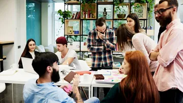 Drei Leute sitzen an einem Holztisch und reden. Eine trägt ein gestreiftes Hemd, eine ein rotes T-Shirt und eine ein blaues Hemd. Auf dem Tisch liegt ein Notizbuch. Im Hintergrund gibt es Bücherregale und bunte Wände.