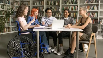 Fünf junge Leute sitzen in einer Bibliothek am Tisch und lernen zusammen.