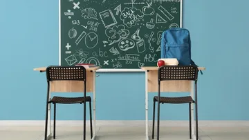 Ein leeres Klassenzimmer mit einem Tisch und zwei Stühlen vor einer beschriebenen Tafel. Ein Rucksack, ein Apfel und Bücher liegen auf dem Tisch. Die Wände sind blau, ein Fenster lässt Licht herein.