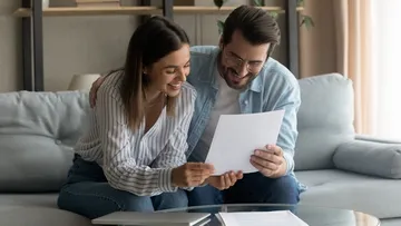 Ein Paar sitzt auf einem Sofa und schaut auf ein Blatt Papier. Die Frau trägt eine gestreifte Bluse und Jeans, der Mann eine blaue Jacke und Jeans. Auf dem Tisch liegen ein Laptop und Papiere. Im Hintergrund sieht man Regale mit Büchern und eine Pflanze.