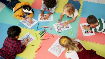Kinder malen Schmetterlingsbilder. Dazu benutzen Buntstifte und Filzstifte. Sie liegen auf blauen, rosa und gelben Schaumstoffmatten und tragen bunte Kleidung.
