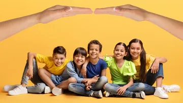 Fünf lachende Kinder sitzen zusammen vor einem gelben Hintergrund, über ihnen halten zwei Hände wie ein schützendes Dach.